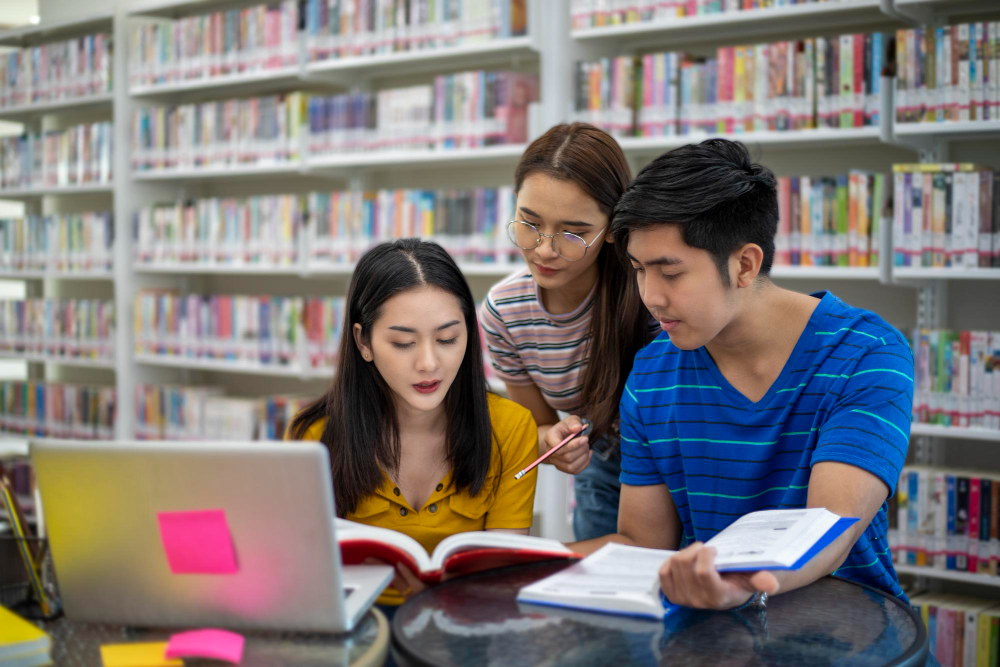 Mahasiswa aktif belajar bersama di perpustakaan dengan buku dan laptop terbuka.