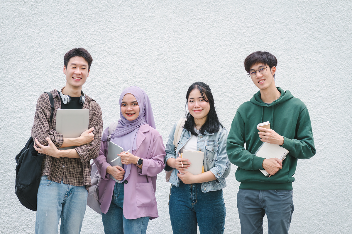 Mahasiswa baru tersenyum sambil membawa buku dan laptop di depan dinding putih.